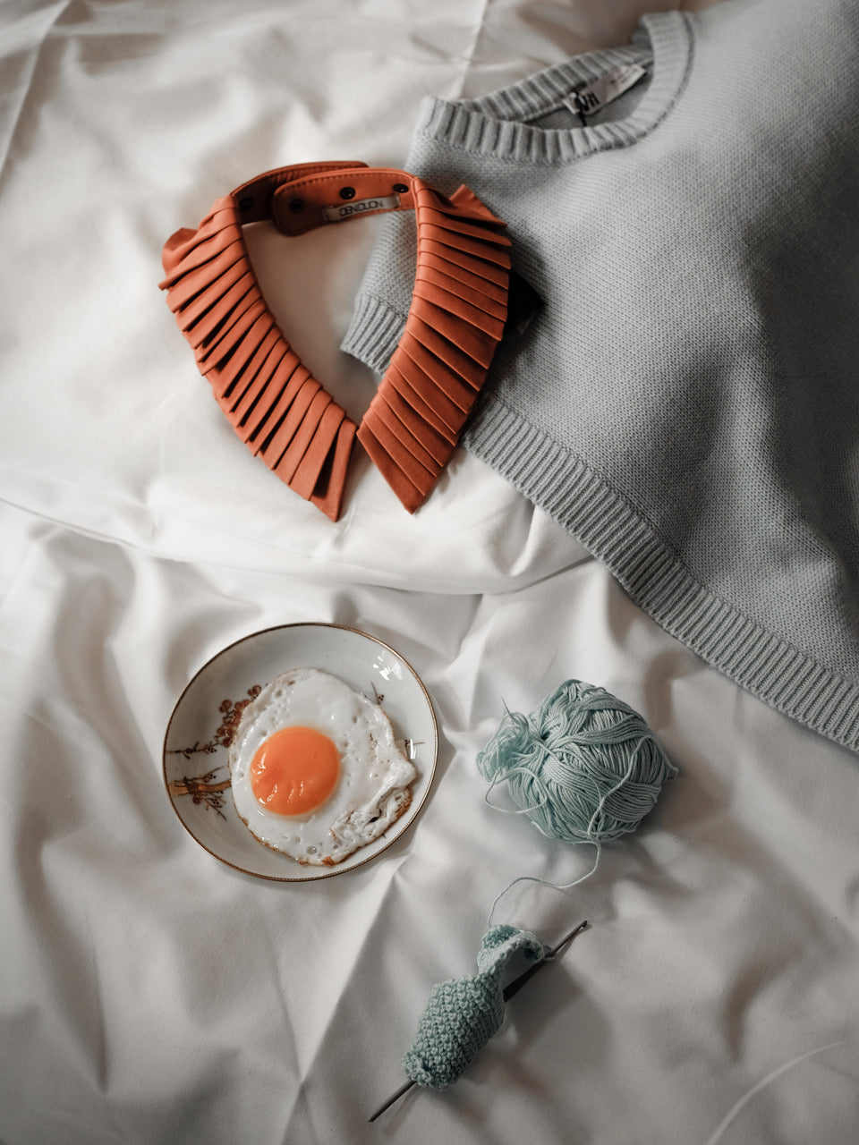 Denduon pleated leather collar in blooming dahlia styled on a hotel bed with a slipover, fried egg and yarn